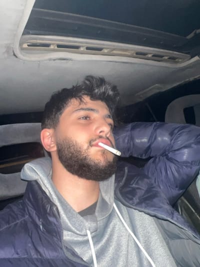 A person with dark hair and a beard is holding a cigarette in their mouth. They're wearing a gray hoodie and a dark blue puffer jacket. The background is blurry but appears to be an interior space, possibly inside a vehicle or a confined area with a white wall or panel visible. The image is rotated, with the person appearing to be lying down on their side.