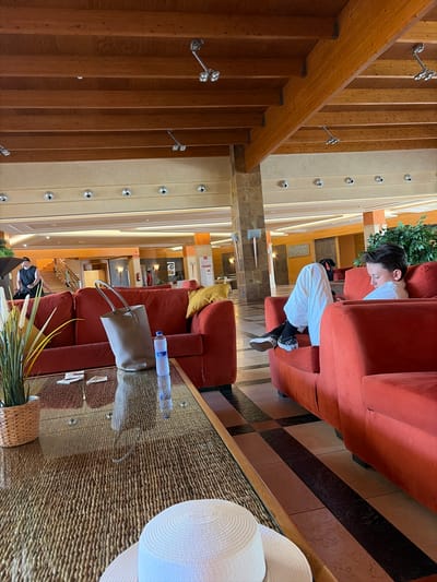 The image depicts the lobby of a hotel in Antigua, Spain. In the foreground, a glass-topped coffee table with a woven base is visible, displaying a potted plant, a grey tote bag, and a bottle of water. A white sun hat rests on the edge of the table. In the mid-ground, a person is seated on a red sofa, looking down at their phone. Further back, another person stands near a staircase. The lobby features red sofas, wooden ceiling beams, and tiled flooring. The overall scene suggests a relaxed atmosphere within the hotel.