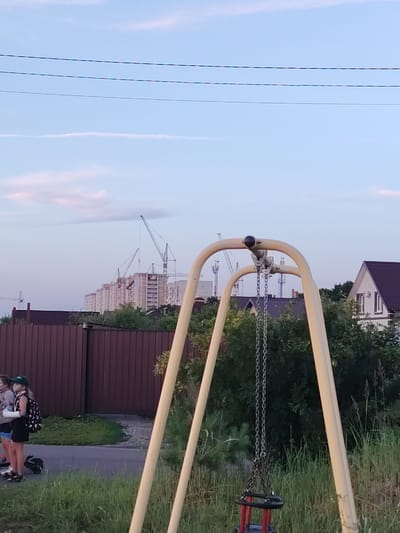 The image depicts an outdoor scene with a playground swing set in the foreground. The swing set has a yellow metal frame and chains suspending a black and red seat. Behind the swing set, a brown wooden fence runs horizontally across the mid-ground. Beyond the fence, there are several tall construction cranes and unfinished buildings, indicating ongoing development in the area. The sky above is a light blue with some wispy clouds and power lines stretched across it. To the left of the fence, a group of three individuals are standing on a paved path, two of whom appear to be young girls. One girl is wearing a black dress with a white polka dot backpack, and the other is wearing a green cap. The environment appears to be a suburban or semi-rural area on the outskirts of the city of Krutye Vyselki, Russia.