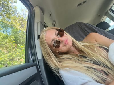 A young blonde woman with long hair and sunglasses is lying in the back seat of a vehicle. She is wearing a white top and a patterned top underneath. Through the open window, a blurry view of green foliage and trees is visible, suggesting the vehicle is in motion. The interior of the car is also visible, with grey and black upholstery. The location is identified as Dražin Vrt, Montenegro.
