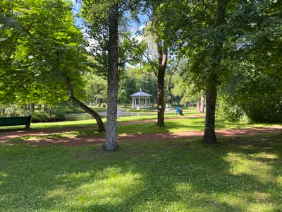 This media file captures a serene park scene in Narva-Jõesuu, Estonia, on a bright, sunny day. The image is dominated by lush green trees, casting dappled shadows on a reddish-brown path that winds through the park. In the foreground, a verdant lawn is dotted with small white wildflowers. To the left, a vibrant green park bench sits near the edge of the path, offering a place to rest.
Further into the park, a picturesque white gazebo stands by a tranquil body of water, reflecting the clear blue sky above. The gazebo appears to be a central feature of the landscape. In the middle ground, a person is visible near the gazebo, tending to what looks like a water fountain or a garden feature. They are dressed in blue, and a blue trash bin is nearby.
The scene evokes a sense of peace and natural beauty, characteristic of a leisurely day in a well-maintained park. The sunlight suggests it is likely midday or early afternoon. The presence of mature trees and the well-kept grounds indicate a public park or garden, possibly a popular spot for relaxation and recreation. No visible text is present in the image.