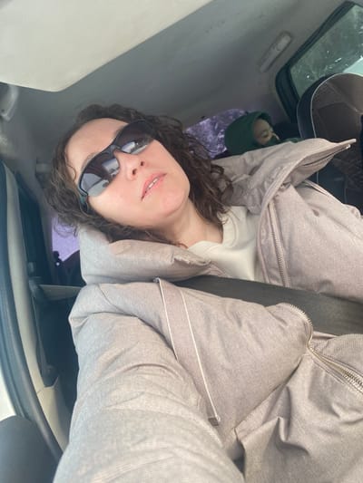 The scene is situated inside a vehicle, reportedly within the city of Ошья, Russia. An adult female is prominently featured in the foreground, occupying the front passenger seat, angled upward as if taking a selfie. She has dark, wavy hair and is wearing dark-tinted sports-style sunglasses, a light-colored crew-neck top, and a light-mauve or taupe-colored quilted winter jacket with a visible zipper closure. A dark-colored seatbelt diagonally crosses her torso. In the background, behind the woman and to her right, a child is partially visible in a dark-colored car seat, wearing a dark green hooded garment. The vehicle's interior ceiling is light-colored. Through the rear window, blurred outdoor elements, consistent with vegetation under daylight conditions, are discernible.