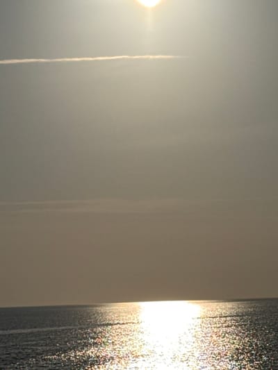 The image presents an open seascape and sky, located in Palaiopoli, Greece. The bright, circular sun is centered in the upper third of the frame, casting a prominent, sparkling reflection across the water's surface from the horizon line towards the foreground. The water appears dark with numerous points of light from the sun's reflection. A distinct, thin, linear contrail is visible horizontally in the upper left quadrant of the sky. The sky itself is largely featureless and hazy, displaying a uniform light gray tone. The horizon line bisects the frame horizontally, separating the dark expanse of water from the luminous sky. No individuals, watercraft, or landmasses are observable.