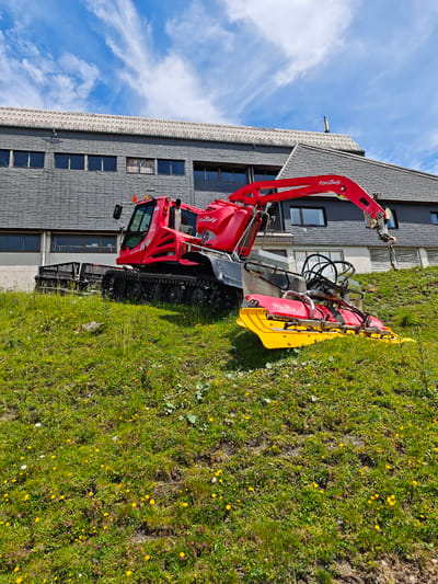 This is a wide, low-angle, exterior, daylight, medium shot of a red and yellow PistenBully snow groomer positioned on a grassy slope dotted with small yellow and purple wildflowers. The vehicle is a multi-purpose utility vehicle with a cab, tracks, a hydraulic arm with a hook, and a tiller attachment. The arm is extended and angled downwards. The PistenBully is in front of a large, multi-story building with a dark grey, sloped roof and a dark grey, horizontal siding facade with multiple windows. The building appears to be a lodge or support structure, possibly at a ski resort. The sky is bright blue with scattered white, wispy clouds. The image does not contain any people. The scene is likely in Kirchberg in Tirol, Austria, based on the presence of a PistenBully, which is commonly used at ski resorts.