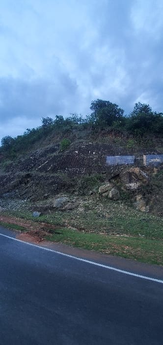 The image displays a landscape scene featuring a road running alongside a rocky, vegetated hillside under an overcast sky, likely in Mua ward, Kenya.
The upper portion of the image shows a light blue sky with scattered, diffused greyish clouds, indicating either an overcast day, early morning, or late afternoon. To the left, a steep, dark grey rocky hillside dominates the frame. The rock face exhibits visible striations and is sparsely covered with green bushes and darker trees, especially towards its crest. Two rectangular, dark, and somewhat reflective panels are affixed to the hillside, appearing weathered or damaged, rendering any potential text unreadable. Lighter patches are also visible on the rock surface.
To the right of the hillside, a paved, dark grey road is visible, marked by a distinct white line along its edge. The road surface appears damp or wet, suggesting recent rainfall or high humidity. A narrow strip of green grass and reddish-brown soil separates the road from the base of the hillside. No people or active events are present, conveying a tranquil and natural setting. The overall mood is subdued due to the cloudy conditions.