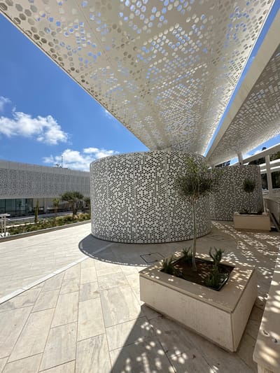 The image depicts an outdoor architectural scene, likely a public space or modern building exterior, characterized by clean lines and geometric patterns. The dominant features are a large, curved building facade with a distinctive perforated white paneling, creating a honeycomb-like effect. Above, a similarly patterned white canopy extends, casting intricate shadows on the paved ground. The ground itself is laid with large, light-colored rectangular tiles. In the foreground, a rectangular planter box contains a small, pruned tree and some greenery, with more planters visible further back. Another cylindrical structure, also with the perforated white paneling, is visible to the right, along with a hint of a modern building in the background with similar patterned elements. The sky is a bright blue with scattered white clouds, suggesting a sunny day. The overall aesthetic is modern, minimalist, and designed to incorporate natural light and shadow play. This architectural style and environment are consistent with the modern developments found in the city of Paphos Municipality, Cyprus, particularly in areas focused on contemporary design and public spaces. There are no people visible in the image.