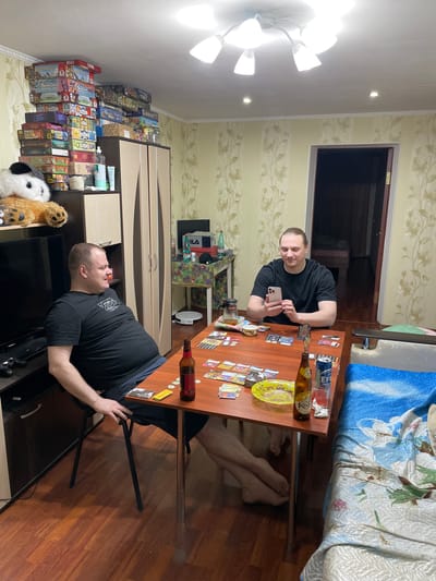 Two men are playing a board game at a table in a room. One man, seated on the left, is wearing a black t-shirt and dark shorts, and is looking to his right. The other man, seated on the right, is wearing a black t-shirt and is looking down at his phone. The table is covered in board game pieces, cards, and bottles of drinks. There is a bed on the right side of the room, with a blue and white patterned bedspread. A large stack of board game boxes is visible on a shelf to the left of the frame. The room has yellow floral wallpaper and a wooden floor. The overall scene suggests a casual gathering in a residential setting. This image was taken in Novocherkassk, Russia.
