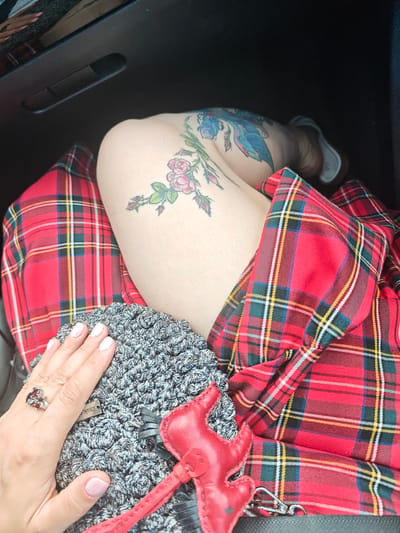 In a medium shot, a person's left leg and hand are visible, resting on a grey crocheted item. The leg is adorned with tattoos of flowers and a bird, and is covered in part by a red tartan fabric. The hand has light pink nail polish and is wearing a silver ring with red stones. The crocheted item has a red leather zipper pull in the shape of a devil. The scene appears to be indoors, possibly in a car given the dark interior visible in the background. The lighting suggests daytime. There is a small black tag with white text on the crocheted item, which is partially obscured but appears to read "Zink."