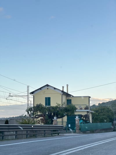 The image depicts a pale yellow, two-story house situated in Pedaso, Italy. The house features a gabled roof with dark trim and two light-colored chimneys. A prominent window on the front facade is fitted with closed dark green louvered shutters. To the right, an extension of the house includes a small balcony with a metal railing, adorned with potted plants, and a smaller dark green door or shuttered window.

In the foreground, an asphalt road with a double solid white center line and a solid white edge line runs horizontally. A metal guardrail runs parallel to the road, partially obscuring the lower portion of the property. The property entrance features a dark green metal gate flanked by two brick pillars, each topped with a white urn containing plants. A large, sprawling tree with dark green foliage and visible branches partially covers the house and entrance area. Additional green fencing and vegetation are present on the right side of the property.

In the background, beyond the house, rolling hills covered with vegetation are visible. To the left of the house, overhead railway lines and a metal catenary mast are present. The sky above is clear and light blue with faint clouds near the horizon. No individuals are present in the scene, and no explicit actions are depicted. The lighting indicates daylight conditions, possibly late afternoon or early morning.