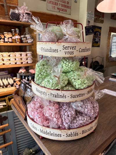 The image shows a tiered display of confectionery items in clear plastic bags, housed in red metal baskets with decorative rims. The rims are labeled with French text, indicating the types of sweets. The top tier contains purple and green sweets, labeled "Friandises - Servez vous - Confiseries." The middle tier has green sweets, labeled "Nougats - Pralinés - Sucettes - Bonbons." The bottom tier holds pink and purple sweets, labeled "Cakes - Madeleines - Sablés - Biscuits - Cookies."
The display is situated on a wooden counter in what appears to be a shop. In the background, shelves are visible, stocked with jars, bottles, and other packaged goods. Several signs with French text are also mounted on the wall behind the display. The overall setting suggests a French-style patisserie or confiserie. The image is well-lit, with the focus on the confectionery display. There are no people visible in the image.