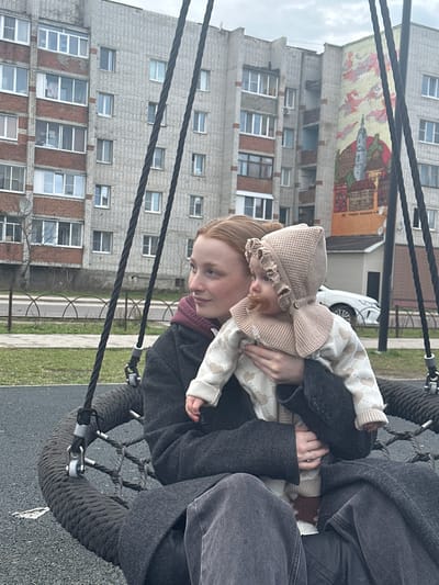 A woman holds a baby, wearing a light-colored bonnet with a large bow and a pacifier, in a swing at a playground in Pokrov, Russia. Behind them are multi-story brick apartment buildings, one of which has a large mural on the top. The sky is overcast. The woman is wearing a dark coat, and the baby is wearing a light-colored sweater with hearts. The playground has a black rope swing and a gray surface. A white car is visible in the background, near a small, light-colored building.
