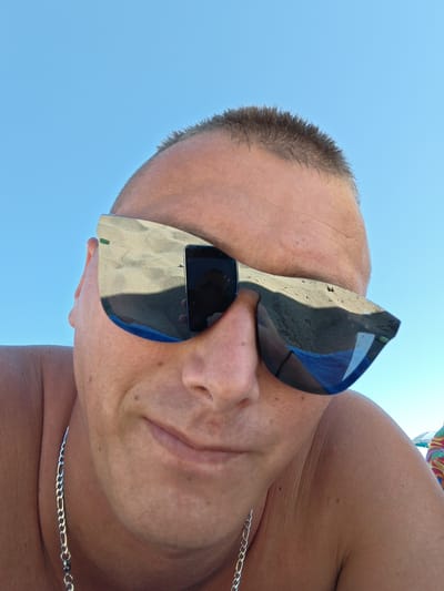 The photograph is a close-up selfie of a fair-skinned man wearing sunglasses, likely taken at a beach in the Gouves Municipal Unit, Greece. The sunglasses reflect the sand and sky, suggesting the location is a beach. The man has short, light brown hair, a chain around his neck, and is shirtless. The sky is clear and blue. A smartphone is visible in the reflection of the sunglasses. The man appears to be holding a device to capture the image.