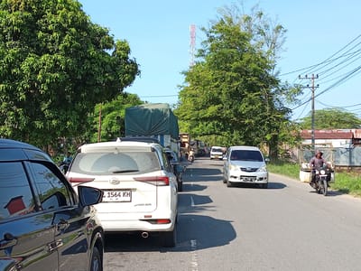 In Blangpanjoe, Indonesia, a traffic jam unfolds on a paved road under a clear blue sky. The foreground is dominated by the side of a black car, reflecting the surrounding environment. Behind it, a white SUV, identified by the license plate "L 1564 KH," is stationary. Further down the road, a silver van is also stopped, and beyond that, a line of trucks, one with a green tarp covering its load. A lone motorcyclist, wearing a hooded jacket, navigates the traffic, carrying bags on the back of his motorcycle. Tall green trees line both sides of the road, and in the distance, a tall communications tower is visible. Utility poles with numerous wires are also present. The scene depicts a typical day with moderate traffic on a road in Blangpanjoe.