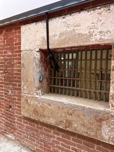 The image shows a close-up, eye-level view of the exterior of a brick building with a barred window. The building appears to be old, with weathered red brick and a stone window frame that shows signs of chipping and decay. A black strap hangs down from a metal fixture above the window, obscuring part of the view through the bars. Inside the window, prison-like bars are visible, and beyond them, a dimly lit interior with what appears to be painted walls is discernible. A round, teal-colored plaque with the number "3" in white is affixed to the stone frame to the left of the window. The ground in front of the building is covered in concrete or a similar material. This scene is located at the former prison "La Model" in Mataró, Spain, which is now a cultural center.