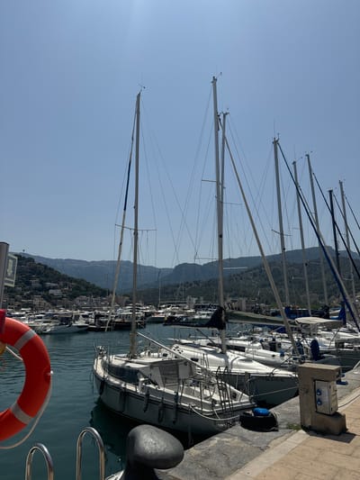 A sunlit harbor scene in El Port, Spain, features a fleet of sailboats moored in turquoise water under a clear blue sky. The foreground is dominated by the side of a white sailboat, its mast reaching high, with a red life preserver hanging on the left. Beyond, numerous other sailboats and smaller vessels are docked. In the background, a densely populated hillside covered in buildings and lush greenery rises towards rolling mountains. A concrete power pedestal with electrical outlets is visible on the right. The image captures a tranquil day at the marina.