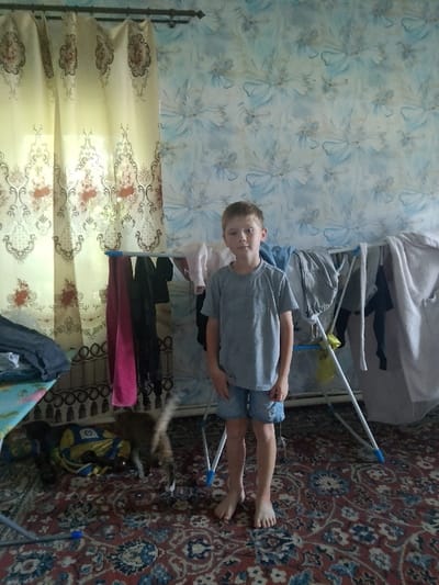 A young Caucasian boy stands barefoot in the center of the frame, facing forward, on a patterned rug. He has short, light brown hair and is wearing a grey t-shirt and blue denim shorts. To his left, a clothes drying rack holds several items of clothing. A cat with tabby markings is visible on the rug near the drying rack, its body angled away from the camera and its tail extended. Behind the boy, the wall is covered with patterned wallpaper. To the left of the boy, a yellow curtain with a floral pattern hangs. The scene appears to be indoors, possibly a residential room in Изыхские Копи, Russia, given the context.