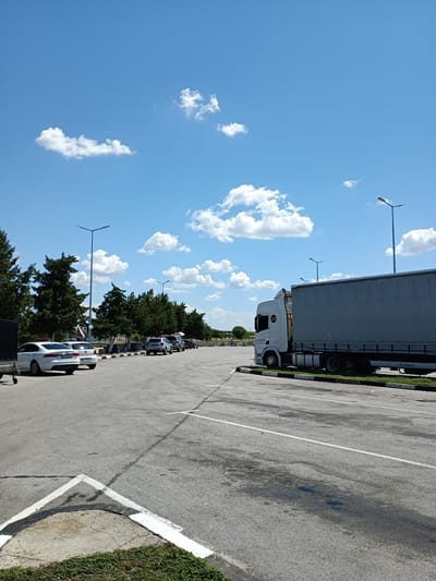 A bright, sunny day is depicted in Radinovo, Bulgaria, with a clear blue sky dotted with fluffy white clouds. The scene is a parking lot, likely at a truck stop or rest area, given the presence of a large semi-trailer truck parked on the right side of the frame. The truck has a white cab and a light gray trailer. In the background, several cars are parked in a row, and more trees are visible. The ground is paved asphalt, with white parking lines and some dark oil stains. Streetlights are visible in the distance, along with more trees and some low vegetation. The perspective suggests the photo was taken from ground level, looking towards the parked vehicles and the horizon.