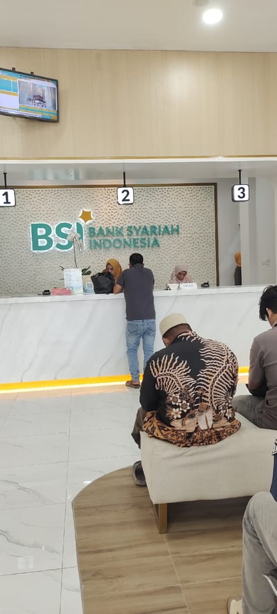 This image depicts the interior of a bank, specifically a branch of Bank Syariah Indonesia (BSI), identified by the prominent logo and name on the wall. The setting is bright and modern, characterized by white marble-patterned counters and flooring, light wooden paneling on the upper walls, and a decorative Islamic geometric pattern behind the counter.
Several individuals are present. In the center, a male customer wearing a dark grey t-shirt and blue jeans stands at the main counter, interacting with a female bank teller. The teller, wearing a brown hijab, is behind the counter, and her nametag reads "MELDA." Another female teller, also in a hijab, is partially visible further down the counter. Queue numbers "1," "2," and "3" are displayed above the counter, indicating service stations.
In the foreground, a man is seated on a light-colored ottoman, facing away from the camera. He is wearing a black shirt with intricate brown and beige batik patterns and a white peci (skullcap). Another person is partially visible sitting beside him. In the background, near counter number 3, another woman wearing a yellow hijab is partially seen.
To the upper left, a wall-mounted flat-screen television displays images, one of which appears to be the Kaaba, along with text including "PT. Bank Syariah Indonesia Tbk" and "KCP Lhoksukon," confirming the specific branch location as Lhoksukon. The overall scene suggests a typical bank operation, with customers being served and others waiting. The presence of hijabs, batik clothing, and the Kaaba on screen highlight the Islamic context of the bank and its location in Indonesia.