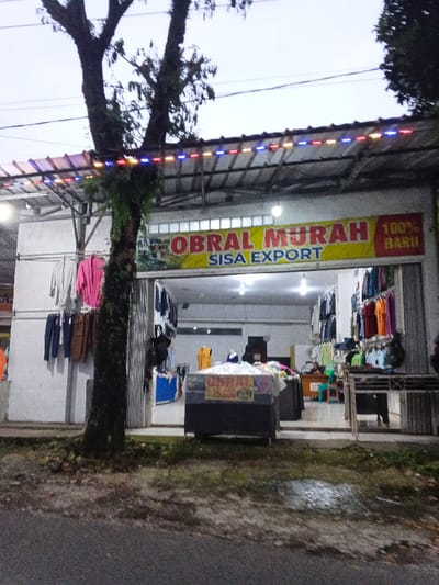 This image depicts a retail clothing store, presented as being located in Phước Hòa, Vietnam, viewed from an outdoor perspective. The scene, captured during dusk or dawn, shows a dark asphalt road with patches of grass and a small wet area in the foreground. A large, dark-barked tree with visible foliage and some moss stands centrally, partially obscuring the store's left side.
The store's white facade features a prominent yellow banner with red text: "OBRAL MURAH SISA EXPORT" and "100% BARU" (Indonesian for 'Cheap Sale Export Surplus' and '100% New'). An awning, comprising corrugated metal supported by a frame, extends over the entrance and is decorated with an illuminated string of red, blue, and yellow LED lights. To the left of the tree, several garments, including white and pink tops and blue and brown pants, are displayed on hangers affixed to the exterior wall.
The store's wide entrance is open, revealing a brightly lit interior with white walls and ceiling lights. Numerous clothing items are arranged on racks along the side walls and deeper within the store, with some mannequins visible. A dark, rectangular display case with a transparent top sits on the pavement directly in front of the entrance, containing merchandise and a white sign with "OBRAL 15.000". A metal rolling shutter is partially visible on the right side of the entrance. Inside, one individual is seated behind a counter in the rear right, and another person is partially discernible near clothing racks on the far right. The store appears operational.