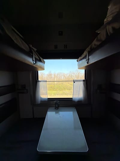 The image shows the interior of a train compartment. There are two bunk beds visible, one above the other. A window with sheer curtains reveals a view of trees and greenery, suggesting a rural landscape. A small, foldable table is attached to the wall on the right side of the window. The lighting is dim, primarily coming from the window. There is no evidence that this is in Lebyazh'e, Russia.