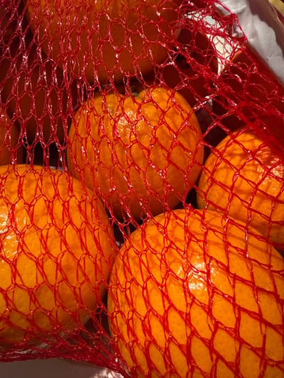 A close-up, top-down view of several ripe oranges, tightly packed within a red mesh bag. The oranges are a vibrant, deep orange color and their textured skins are visible through the diamond-shaped openings of the netting. Some of the oranges appear slightly shiny, suggesting they are fresh and possibly recently washed. The red mesh creates a grid-like pattern over the fruit, with some areas of the oranges obscured by the netting, while others are more clearly visible. The background is out of focus and appears to be a light-colored surface, possibly a grocery bag or a countertop. The lighting suggests an indoor setting, likely a kitchen or a market stall. There is no discernible activity or event taking place, as the image focuses solely on the still life of the oranges. No people are visible, and there are no obvious clues about the time of day, weather, or specific location beyond what might be inferred from the type of product being displayed. No text is visible in the image.