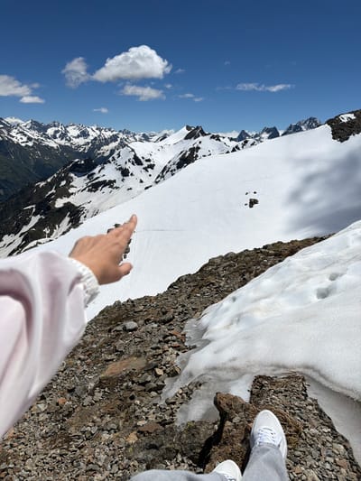 A person's hand, pointing towards a snow-covered mountain range, is visible in the foreground of the image. The person is wearing a light-colored jacket. The background features a vast expanse of snow-capped mountains under a clear blue sky with scattered clouds. The terrain in the foreground consists of rocks and patches of snow. The location is the city of Verkhne-Teberdinskoye rural settlement, Russia.