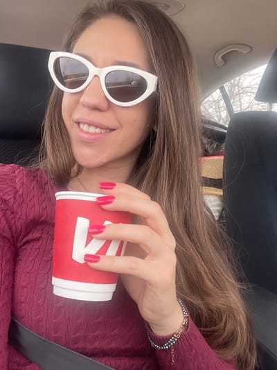 A young woman with long brown hair is inside a vehicle, possibly a car. She is wearing white-rimmed sunglasses and a maroon sweater. She is holding a red and white coffee cup in her right hand, showcasing her red nail polish. A black seatbelt is visible across her torso. The car's interior is visible including the beige car ceiling and a black car seat. The background through the car window appears to show trees, although the focus is soft. Kiosevtsi, Bulgaria, is not discernible from the image.