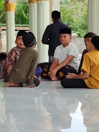 A group of young boys, all wearing traditional peci caps, are sitting on a polished tiled floor. Some are facing each other, engaged in conversation, while others are looking away. The boys are dressed in traditional clothing, including long-sleeved shirts and patterned pants. One boy in the foreground is wearing a green long-sleeved shirt and dark pants, sitting with his legs crossed. Another boy, in the center, wears a white polo shirt and black patterned pants. To his right, a boy in a yellow shirt is also seated. Behind them, more boys and an adult are visible.

The setting appears to be outdoors, possibly a mosque or a traditional building, with large white pillars and lush green foliage in the background. The sunlight suggests it is daytime. The reflective surface of the floor mirrors the figures and the surroundings, adding a sense of depth. The overall mood seems calm and communal, suggesting a gathering or a lesson. There is no visible text in the image. The location context is Lancoklancok, Indonesia.