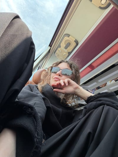 A low-angle close-up photograph captures a person, likely a young woman, inside what appears to be a bus or train in Hải Phòng, Vietnam. The subject is seen from a perspective looking upwards, suggesting they are reclining or positioned low in their seat. She has curly brown hair and wears dark sunglasses, which reflect the bright, cloudy sky outside. Her right hand is raised to her face, partially obscuring her mouth and chin.
She is dressed in loose-fitting black clothing, possibly a hoodie or top and trousers, with visible distressed or frayed edges on the sleeves. Behind her head, a light brown or tan object, possibly a seat headrest, is visible. The interior of the vehicle features overhead panels with integrated lights and air vents, colored in muted tones of cream and burgundy. A large window on the left side of the frame shows a bright, partially cloudy daytime sky. The overall impression is a casual, relaxed moment of travel. No visible text is present in the image.