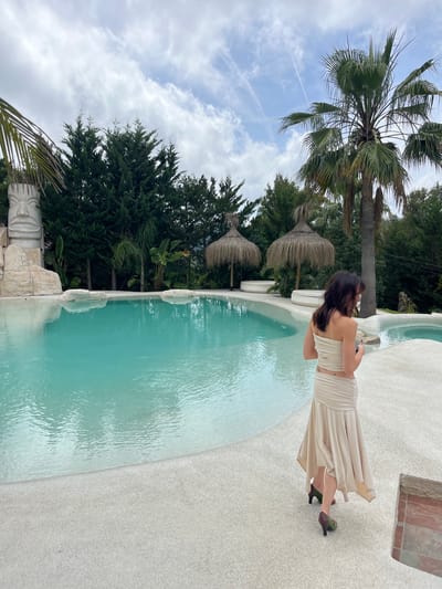 In the city of Can Güell, Spain, a woman stands near a turquoise swimming pool. She is wearing a beige strapless top and a matching flowing skirt, with green and brown heeled shoes. The pool is surrounded by a white pebbled deck, and in the background, lush green trees and palm trees create a tropical ambiance. Two thatched umbrellas stand near white circular lounge beds. To the left, a large stone tiki statue is visible, carved into a rocky outcropping. The sky above is partly cloudy with hints of blue.