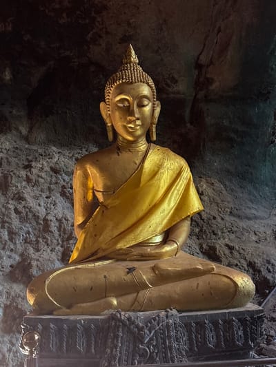 A golden Buddha statue is centrally positioned within a cave in Ban Hin Sam Kon, Thailand. The statue is depicted seated in a cross-legged meditative posture, with its hands resting in its lap. Its surface is golden-colored, featuring a flame-like ushnisha, tightly curled hair, closed eyes, and elongated earlobes. A draped, shiny golden fabric covers the statue's left shoulder and torso. Minor dark smudges and surface wear are visible on the statue, particularly on its legs. It rests upon a multi-tiered, dark-colored base adorned with intricate, dark relief carvings. The background consists of dark, textured rock walls, indicating a natural cave environment, illuminated by an unseen light source that highlights the statue.