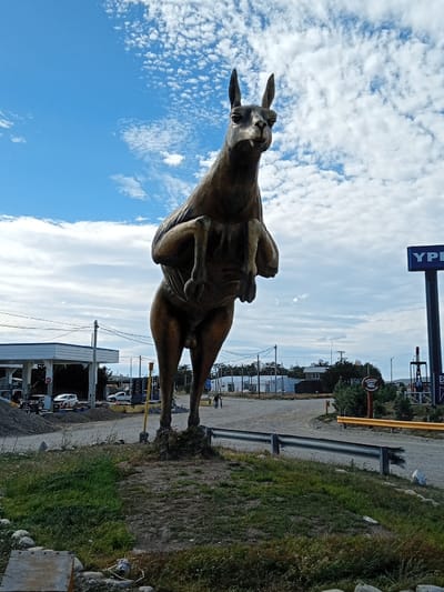 A large, bronze-colored kangaroo statue stands on a grassy mound. It's positioned in front of a gas station or rest stop with a "YPF" sign. The statue is in a rearing position, its front legs tucked towards its chest. The sky is blue with scattered white clouds. A black metal guardrail encircles the base of the mound. In the background, there are buildings and parked cars, suggesting a roadside stop.