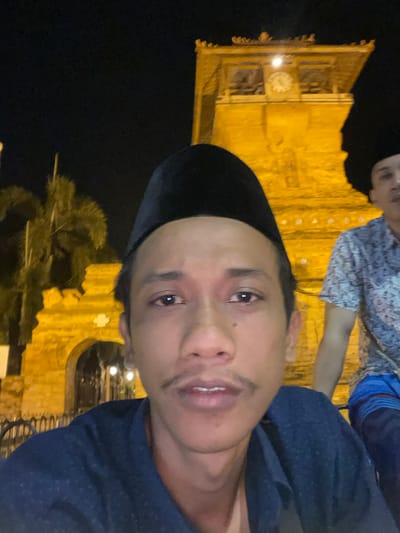 An outdoor, nighttime photograph captures two adult males with the brightly illuminated clock tower of the Great Mosque of Kauman in Yogyakarta, Indonesia, as a prominent background element.

In the immediate foreground, a male, possibly of Southeast Asian ethnicity, is positioned centrally, looking directly at the camera with a neutral expression. He wears a black songkok (traditional cap) and a dark blue collared shirt with a subtle dot pattern. He has a thin mustache and short facial hair below his lower lip. His dark hair is visible under the songkok.

To the right, partially obscured by the foreground subject, a second male is visible from the head to the upper torso. He wears a light-colored, short-sleeved shirt with a dark pattern and blue patterned shorts. A dark head covering, similar to a songkok, is partially visible on his head.

The background features the stone clock tower of the Great Mosque, brightly lit with yellow artificial light. A clock face is visible near the top of the tower, along with ornate architectural details and arched openings. To the left of the tower, a segment of another similarly illuminated stone structure with an arched entrance is visible. Dark silhouettes of palm trees are present against the night sky on the far left. The sky is dark, indicating night.