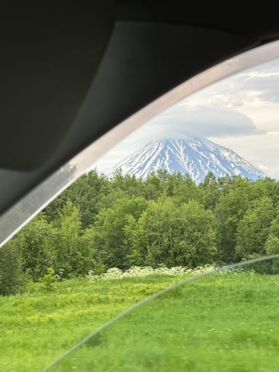 The image shows a view from inside a vehicle, looking out at a snow-capped mountain and a forest. The foreground is dominated by the dark interior of the car, with a section of the windshield and side window visible. A bright, diagonal line suggests the edge of a window frame or a reflection. Beyond this, a lush green field stretches out, dotted with small white wildflowers in the middle ground. Behind the field, a dense forest of green trees rises up. Peeking above the treeline is a majestic, snow-covered mountain with a cloud swirling around its peak. The sky is overcast with a mix of grey and white clouds. The scene depicts a natural landscape, likely outside of a populated area, and does not contain any people or specific urban elements that would help pinpoint a location in KETKINO, Russia.