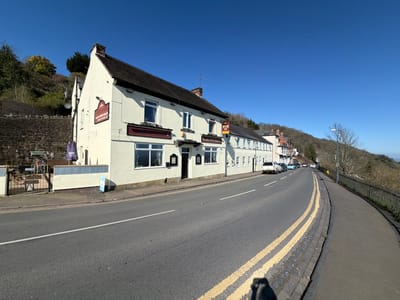 The image depicts The Fleece Inn and Malvern Hills Hotel, located on The Wyche in Malvern, Worcestershire, United Kingdom, under a clear blue sky. The primary subjects are a contiguous cream-painted, two-story building complex on the left side of a curving asphalt road. This structure features a dark tiled roof, a brick chimney, multiple white-framed windows, and a dark main entrance door. Signage prominently displays "The Fleece Inn" on a projecting board and "Malvern Hills Hotel" beneath ground-floor windows.

To the left of the main building, an outdoor patio area with tables, chairs, and a folded purple parasol is visible, enclosed by a low cream-colored wall and a black metal gate. Behind this area and the main building, a steep, vegetated hillside rises, incorporating a stone retaining wall. The asphalt road shows a single white lane marking and distinct double yellow lines along the right curb. A paved sidewalk runs along the right side of the road, separated from a descending, tree-covered hillside by a black metal fence. A white street lamp post is positioned further down the road on the right. Several parked vehicles are visible along the left side of the road adjacent to the buildings. The majority of deciduous trees on the hillsides are bare, indicating a late winter or early spring period. No individuals are present within the frame.