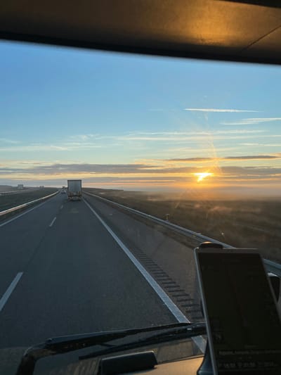 A highway stretches towards a golden sunrise, visible through the windshield. White contrails streak the blue sky above. A large truck moves in the distance on the same lane. The road surface is dark and marked with white lane dividers. A navigation device is attached to the dashboard, displaying time data. The scene captures a journey during the early hours, with the sun illuminating the road and sky.