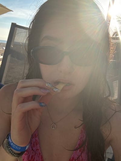 A person wearing sunglasses is eating something fried, possibly a French fry. Their nails are decorated in a unique pattern with shiny embellishments. They wear a necklace with a circular pendant and multiple bracelets, including a blue wristband with text. The sun is shining brightly. They are at an outdoor location.