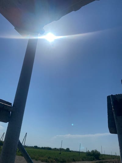 This image captures a bright, sunny day in Ranchpar, Armenia. The sky is a clear, vibrant blue, with the sun shining intensely, creating a strong lens flare and a visible sunbeam extending across the frame. The scene is dominated by a clear sky, but in the foreground, metal structures, likely part of a fence or industrial equipment, partially obscure the view. These structures are weathered and appear metallic.
To the right of the image, a green field stretches into the distance, lined with a road and dotted with utility poles. The lighting suggests it is daytime, likely mid-morning or early afternoon, given the bright sun and the clear visibility of the landscape. The overall impression is one of a rural or semi-industrial setting under a beautiful, clear sky. There is no visible text in the image.