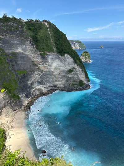 The image shows a coastal scene with a massive cliff face dominating the view. The cliff is layered with grey rock and some vegetation, mainly green shrubs. Below the cliff, there is a small sandy beach with visible waves crashing onto the shore. The water transitions from a deep blue color further out to a light turquoise near the shore, where the wave action is most visible. In the distance, there are a couple of smaller rocky islands with vegetation. The sky is blue with some scattered clouds.
This appears to be a view of Kelingking Beach, located in Bunga Mekar, Indonesia. The steep, rocky cliff face and the characteristic shape of the beach are consistent with this location.