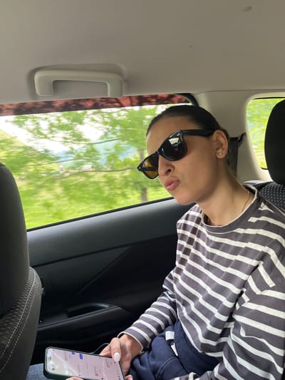 A young woman, wearing sunglasses and a striped long-sleeved shirt, is sitting in the back seat of a car. She is looking to her right and puckering her lips. Her left hand holds a smartphone. Outside the car window, blurred green trees and mountains are visible, suggesting a rural or suburban setting, possibly in or around Achkhoti, Georgia. The interior of the car is dark, with a gray headliner and seats. A grab handle is visible on the ceiling of the car.