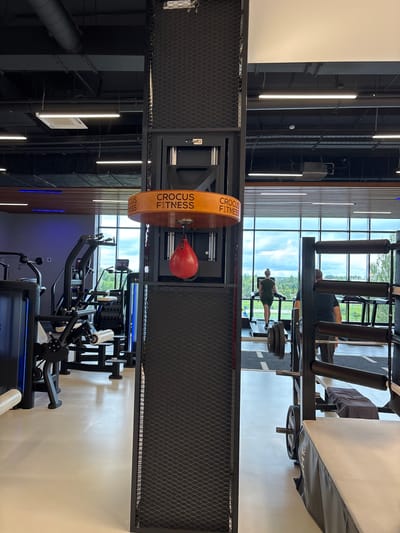 The image shows the interior of a gym, likely located in Monolit, Russia, given the context. In the foreground, a tall, black metal structure with a mesh-like pattern dominates the left side of the frame. Attached to this structure is a red speed bag, suspended from an orange, circular platform bearing the "CROCUS FITNESS" logo. To the left of this setup are various pieces of gym equipment, including a leg press machine.

In the mid-ground, through a large window, two individuals are visible. One person is walking on a treadmill, facing away from the camera. Another person, also seen from the back, is operating a different piece of exercise equipment. The background outside the window suggests a park or outdoor area with trees and a cloudy sky.

The gym environment features a light-colored, polished floor and a modern ceiling with exposed ductwork and fluorescent lighting. The overall impression is of a well-equipped and contemporary fitness facility.