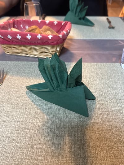 This image depicts a restaurant table setting in Lalden, Switzerland. In the immediate foreground, a dark green paper napkin is intricately folded into a decorative, fan-like arrangement, resting on a light beige textured placemat or tablecloth. To its left, the tines of a silver fork are visible. In the midground, slightly out of focus, a woven wicker basket with a red fabric liner, adorned with white Swiss crosses, contains several pieces of bread. Behind this basket, another dark green folded napkin and a clear drinking glass are discernible. The background shows further out-of-focus elements of a dining area, including portions of another table setting and the upper body of an individual, dressed in dark attire, at the top left. The scene is illuminated by ambient indoor lighting, casting subtle shadows on the table surface.