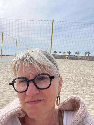 A close-up, tilted, daytime photograph features a Caucasian woman with short, gray hair and black-rimmed glasses. She is positioned in the foreground, and a beige sweater collar is visible on her right side. She also wears earrings.

Behind her, a volleyball net stretches across a sandy beach. Further in the background, a row of palm trees lines the beach, and a building or structure is visible, bearing the inscription "Bar à Cocktails." The sky is overcast and pale.

Based on the presence of the palm trees and the "Bar à Cocktails" sign, the location is likely a beach in the city of Palavas-les-Flots, France.