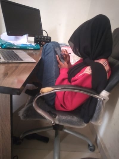 An individual is seated in a black executive-style office chair in an indoor environment. The person wears a black hijab, a red long-sleeved top, and blue jeans; their face is obscured. They hold a dark-colored smartphone in their left hand and operate it with a pink stylus held in their right hand. The office chair has a five-star base with casters, and its seat and back are covered with clear plastic. A small, light-brown, elongated object rests on the right armrest. To the left, a wooden desk supports an open silver laptop, a black computer monitor, a black handbag with gold-colored hardware, a light blue and white "NEXIN" tissue box, and stacked white papers. The walls are light-colored, and the floor is tiled. The specific location within Kanti, Nigeria, cannot be determined from the visual information provided.