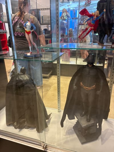 An indoor shot of a glass display case shows two Batman action figures and a Supergirl action figure. The Batman figures are on the bottom shelf, and the Supergirl figure is on the top shelf. A person wearing a dark t-shirt is visible behind the Supergirl figure. The figures are well-lit, and the background is out of focus.