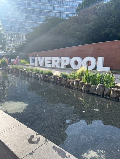 The image shows a public art installation spelling "LIVERPOOL" in large white block letters, set beside a shallow water feature lined with plants and rough stone edging. The letters are positioned in front of a brick wall. A modern building with a grid of windows can be seen on the left. A group of people stand near the installation. The photo appears to be taken at ground level. The location is identifiable as Liverpool, United Kingdom due to the name of the city being displayed in the artwork.