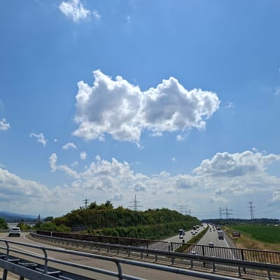 This media file depicts a sunny day with scattered clouds over a German highway. The scene is viewed from an overpass, looking down at a multi-lane highway where vehicles are traveling in both directions. Prominent features include a densely vegetated hillside to the left, dotted with electrical pylons, and a green agricultural field to the right. In the distance, faint outlines of mountains can be seen, and a church spire is visible above the trees near a town. The sky is a vibrant blue, with fluffy white cumulus clouds casting soft shadows. The overall impression is one of a typical day on a European highway, characterized by moderate traffic, infrastructure, and rural scenery. There is no visible text in the image.