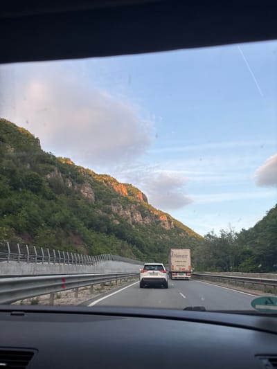 The image, taken from inside a vehicle, shows a view of a highway winding through a mountainous, forested landscape. A white car and a truck with the name "Hamster" printed on the side are visible on the road. The sky is partly cloudy with a contrail visible. The location appears to be near Novo Bardo, Bulgaria, based on the license plate "PB 0118" on the car, which is consistent with Bulgarian registration plates. The mountain slope is covered in dense green vegetation. There's a guardrail along the side of the highway. The image has a slight tilt and obstruction on the left side due to the vehicle's interior.