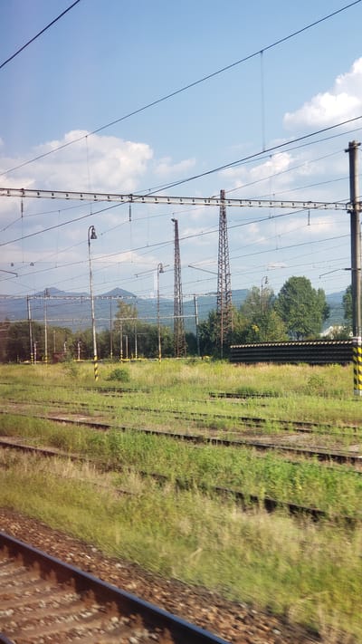 The image depicts a view of train tracks and overhead electrical wires in the city of Martin, Slovakia, likely taken from a moving train. In the foreground, multiple sets of train tracks run parallel to each other, with one set in sharp focus and others receding into the distance. The tracks are partially overgrown with tall, green grass and wildflowers.
In the middle ground, a large grassy area is dotted with trees of varying heights and foliage density. Several tall, slender utility poles with lights are visible, as are two tall, lattice-like metal towers. A long, dark, rectangular structure, possibly a building or a covered platform, is situated behind some of the trees. In the background, a range of distant, hazy mountains forms the horizon under a bright blue sky with scattered white clouds. The overall impression is a semi-rural or industrial area adjacent to the railway.