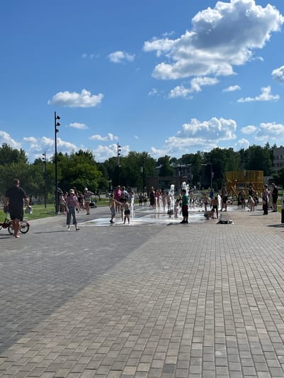 A full, outdoor, eye-level shot shows a crowd of people, many of them children, enjoying a splash pad on a bright, sunny day in Kirovo-Chepetsk, Russia. The splash pad is in the center of the frame, with water jets spraying upwards. Children are running through the water, laughing and playing. Adults are standing around, some watching the children and others interacting with them. The ground around the splash pad is paved with gray interlocking bricks, and a few bicycles are parked nearby. In the background, there are trees, buildings, and a playground structure. The sky is blue with fluffy white clouds. The overall scene is one of joy and recreation.